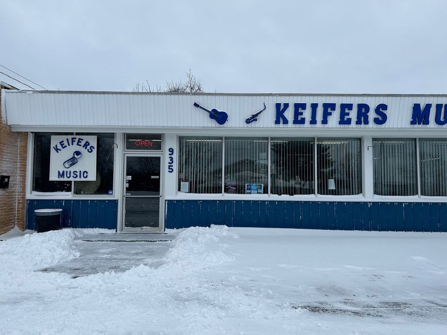 Keifers Music store front in winter. Blue and white sign, snow on the ground, glass windows.