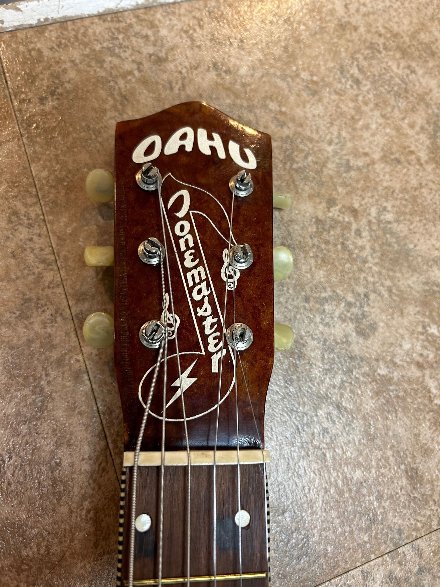 Headstock of an Oahu Tone master guitar with white lettering, brown finish, and six tuning pegs.