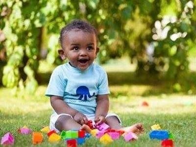Smiling baby in a light blue shirt sitting on grass with colorful toys and flowers in the foreground