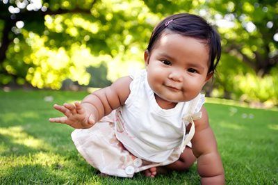 Baby crawling on green grass outdoors, wearing a white outfit and reaching forward with a curious smile