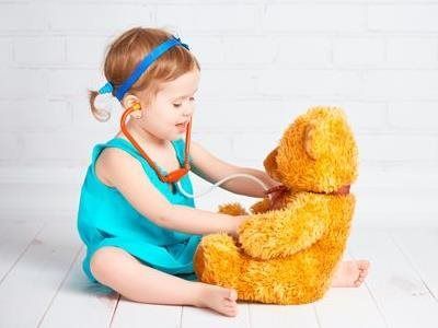 Young child in a turquoise outfit stethoscoping a plush teddy bear on a white floor
