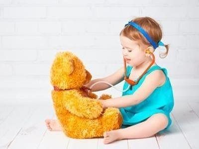 Child in a blue outfit playing with a teddy bear on a white floor, sitting against a white wall