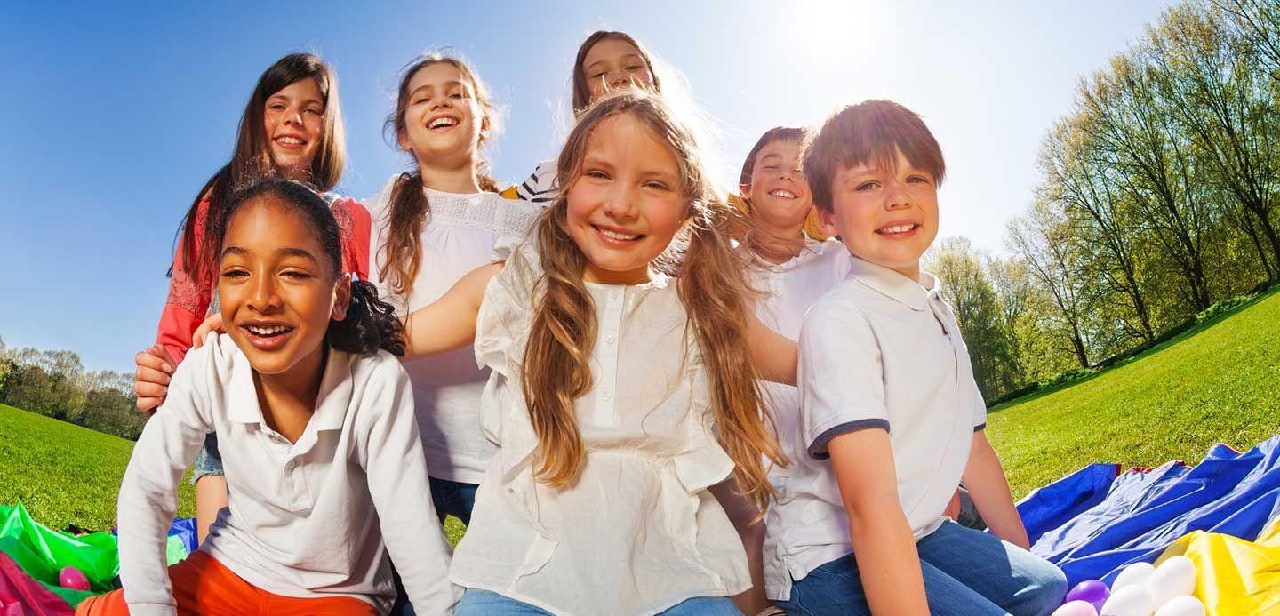 Children smiling on a colorful blanket in a sunny park, with trees and blue sky behind them