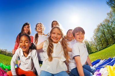 Children smiling and sitting on a colorful blanket in a sunny park with trees and blue sky