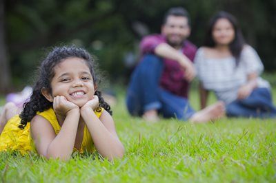 Smiling child lying on grass with hands under chin, with two blurred people sitting in the background.