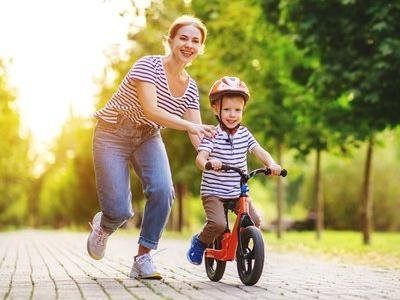 Woman helping child ride a balance bike on a sunlit park path