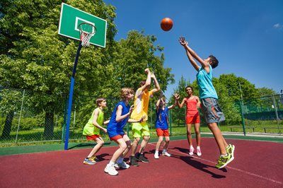 Children playing basketball on an outdoor court, one shooting toward the hoop on a sunny day.