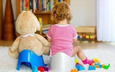 Two children sitting on small chairs, facing bookshelves and toys in a bright playroom.