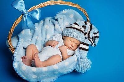 Sleeping baby in a blue plush cradle with a striped knit hat and white pacifier on a blue background