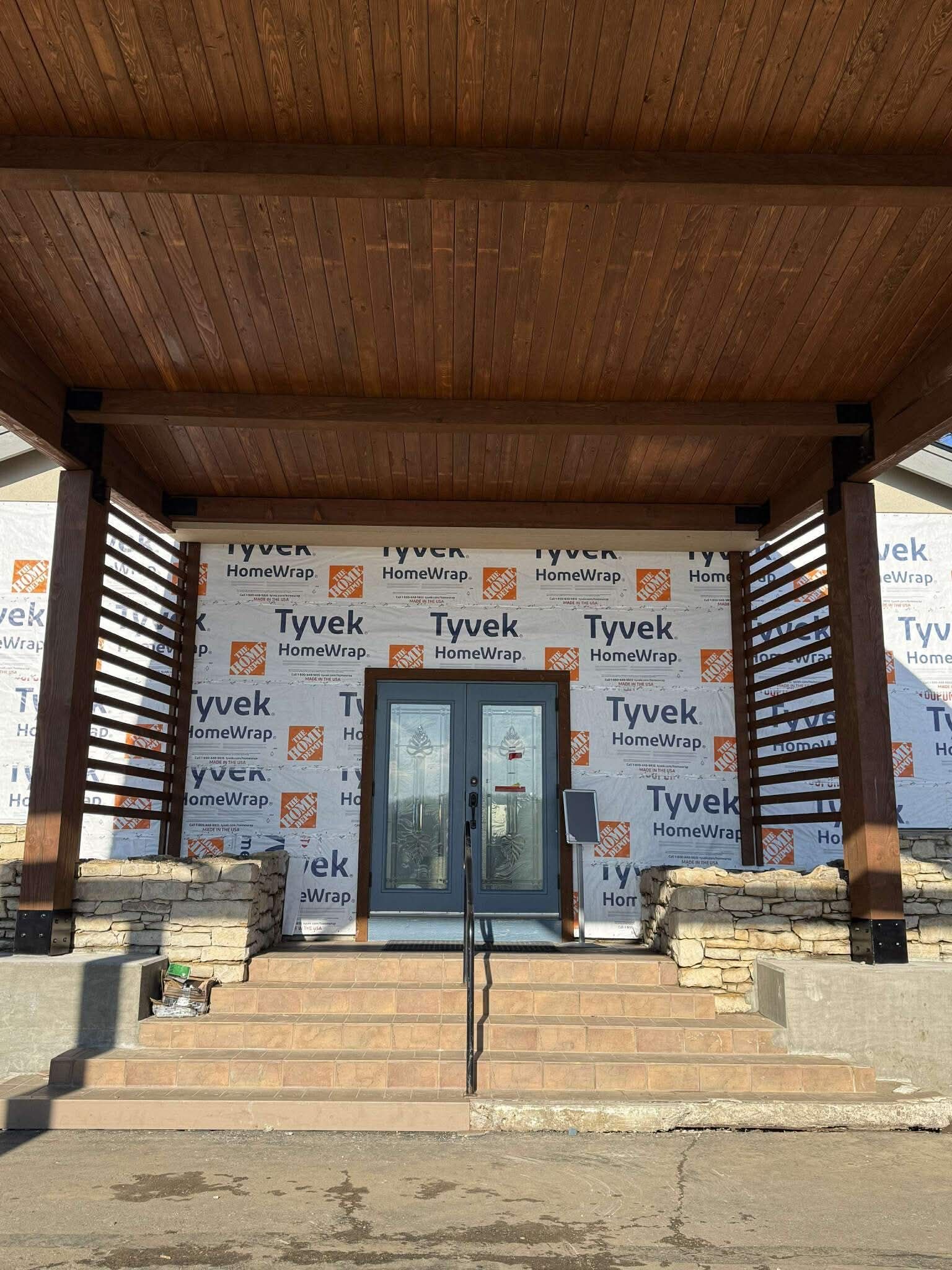 Exterior of building under construction with double doors, under a wooden awning. Stone steps lead up to the entrance.