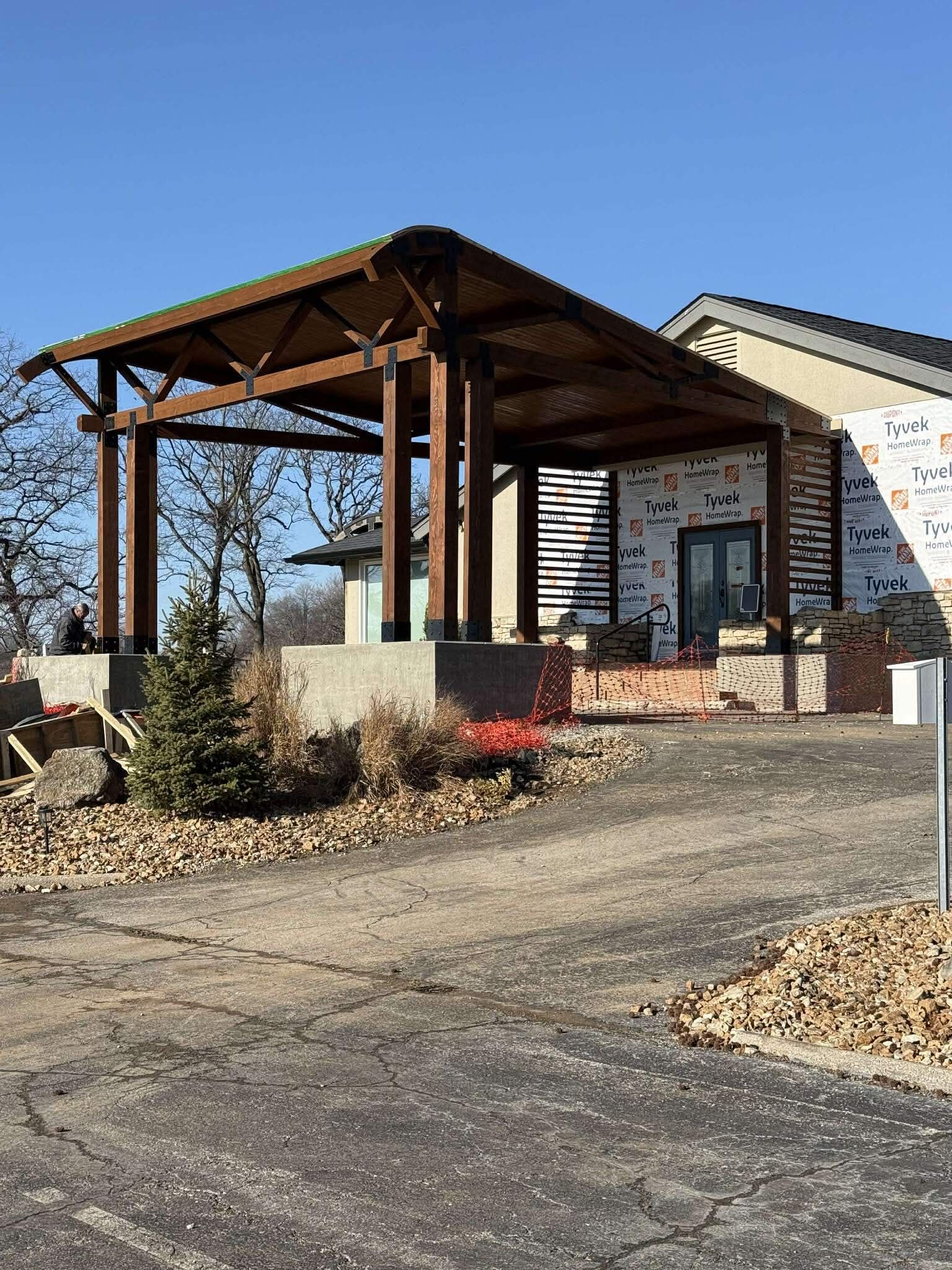Wooden pergola over a concrete patio; small evergreen bushes and flowers in front. Paved driveway, blue sky.