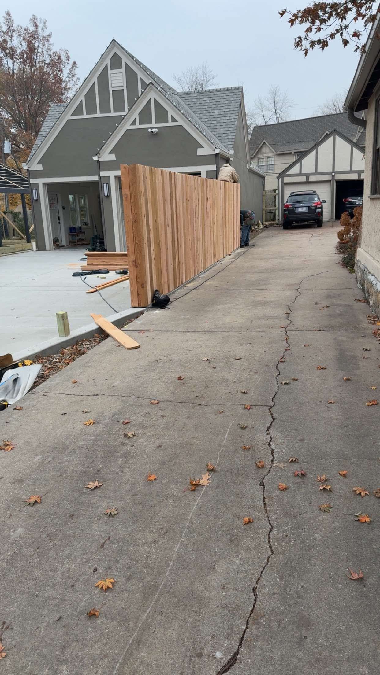 Driveway next to a wooden fence, leading to a house. Gray overcast sky.