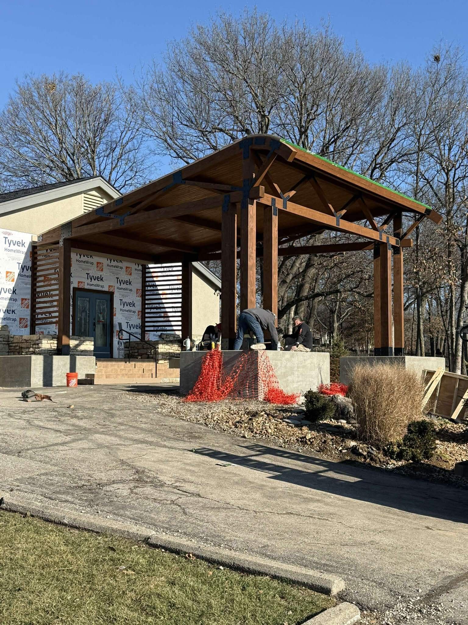 Construction of a covered patio with wood beams. Workers on the concrete base, trees in background, sunny day.