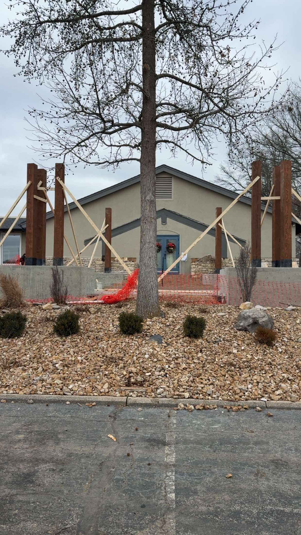 Building with wooden posts and braces, tree in front, mulched landscaping, overcast sky.