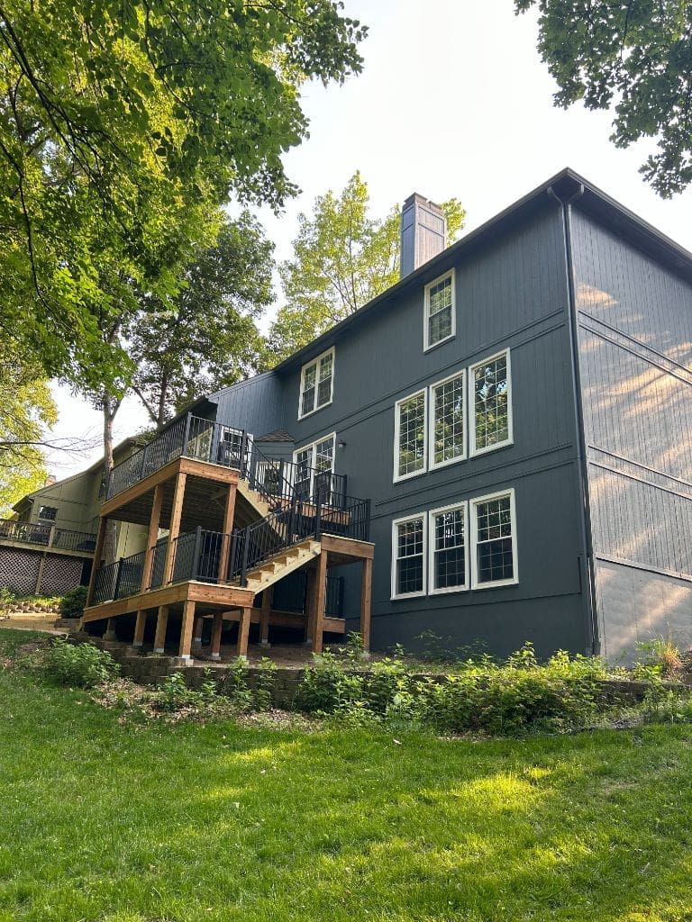 A large black house with a deck and stairs is sitting on top of a lush green hillside.