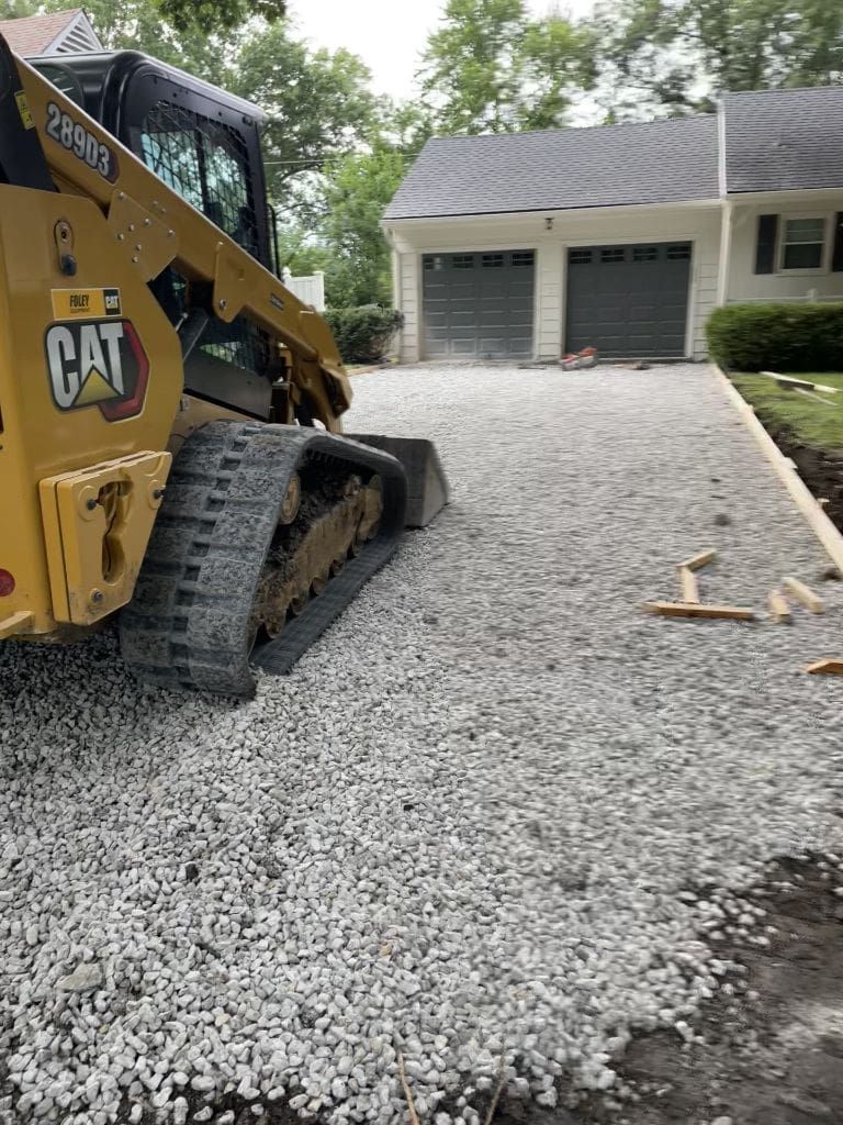 A cat skid steer is working on a gravel driveway in front of a house.