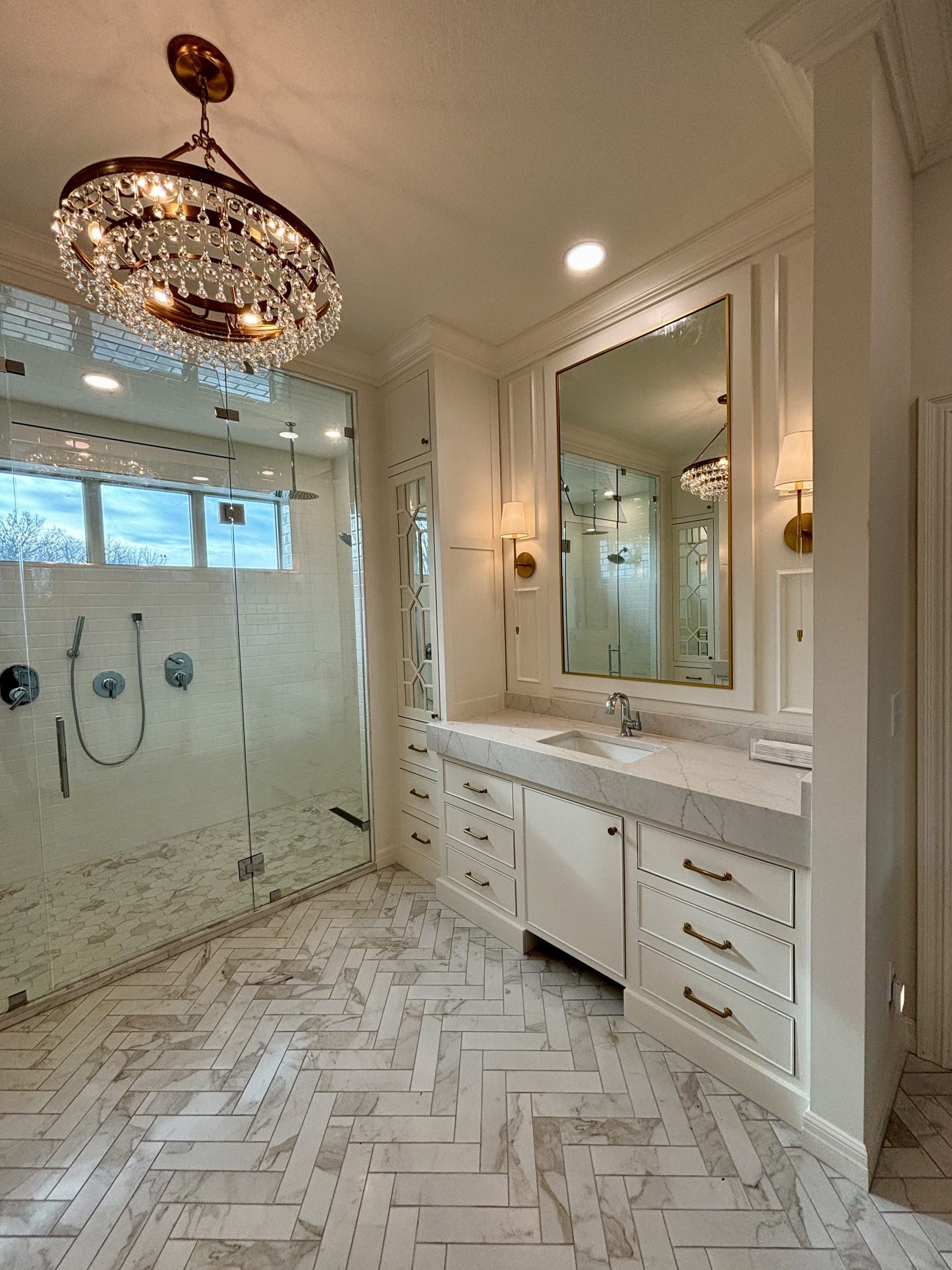 A bathroom with a walk in shower , sink , mirror and chandelier.