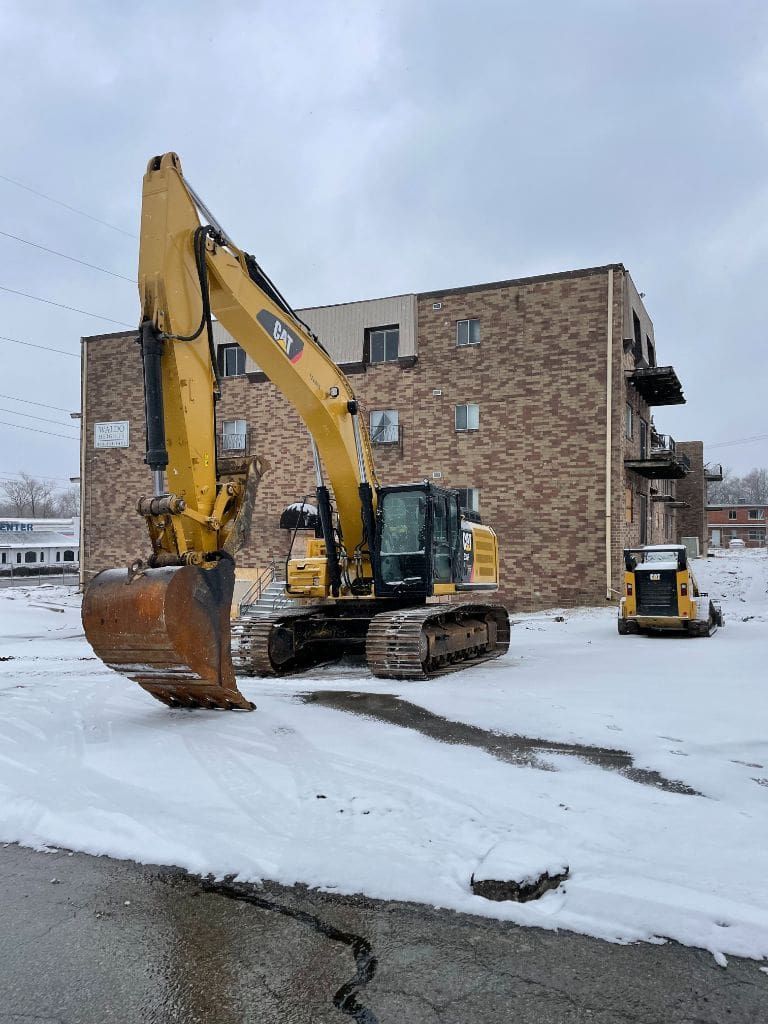 A yellow excavator is parked in the snow in front of a brick building.