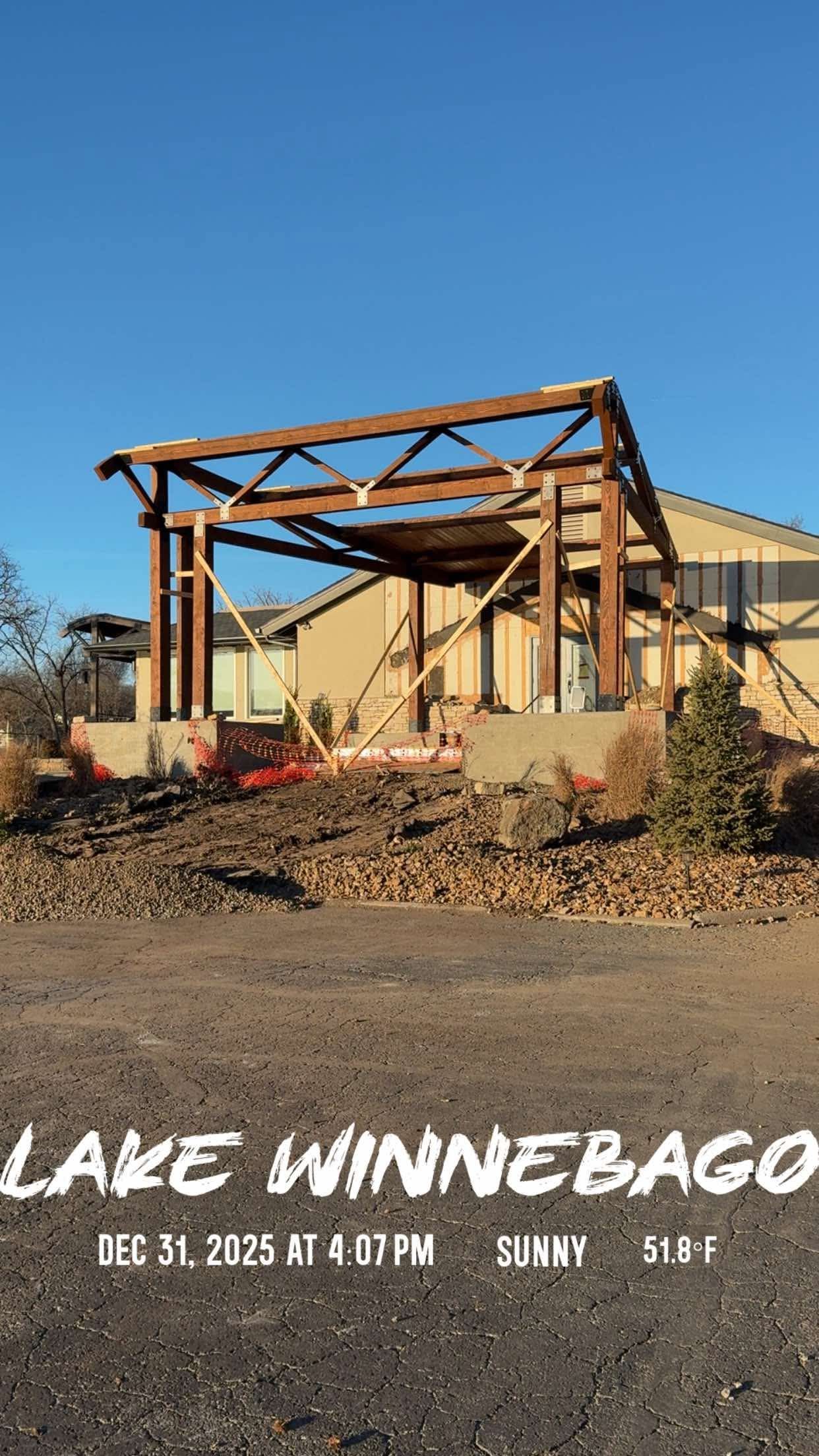 A partially constructed wooden structure in front of a tan building under a clear blue sky.