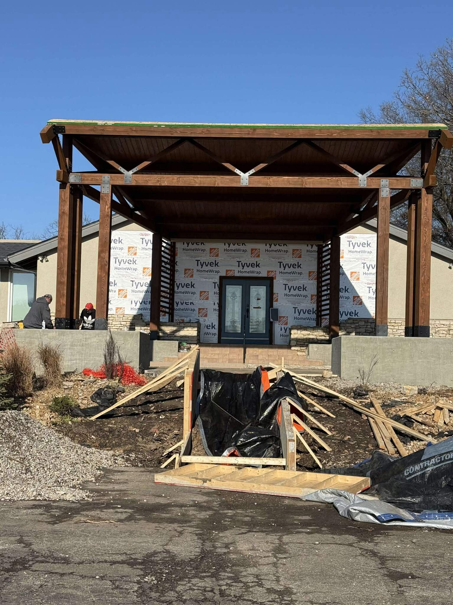 House under construction with pergola over entrance. Two doors, Tyvek wrap, wood and stone facade.
