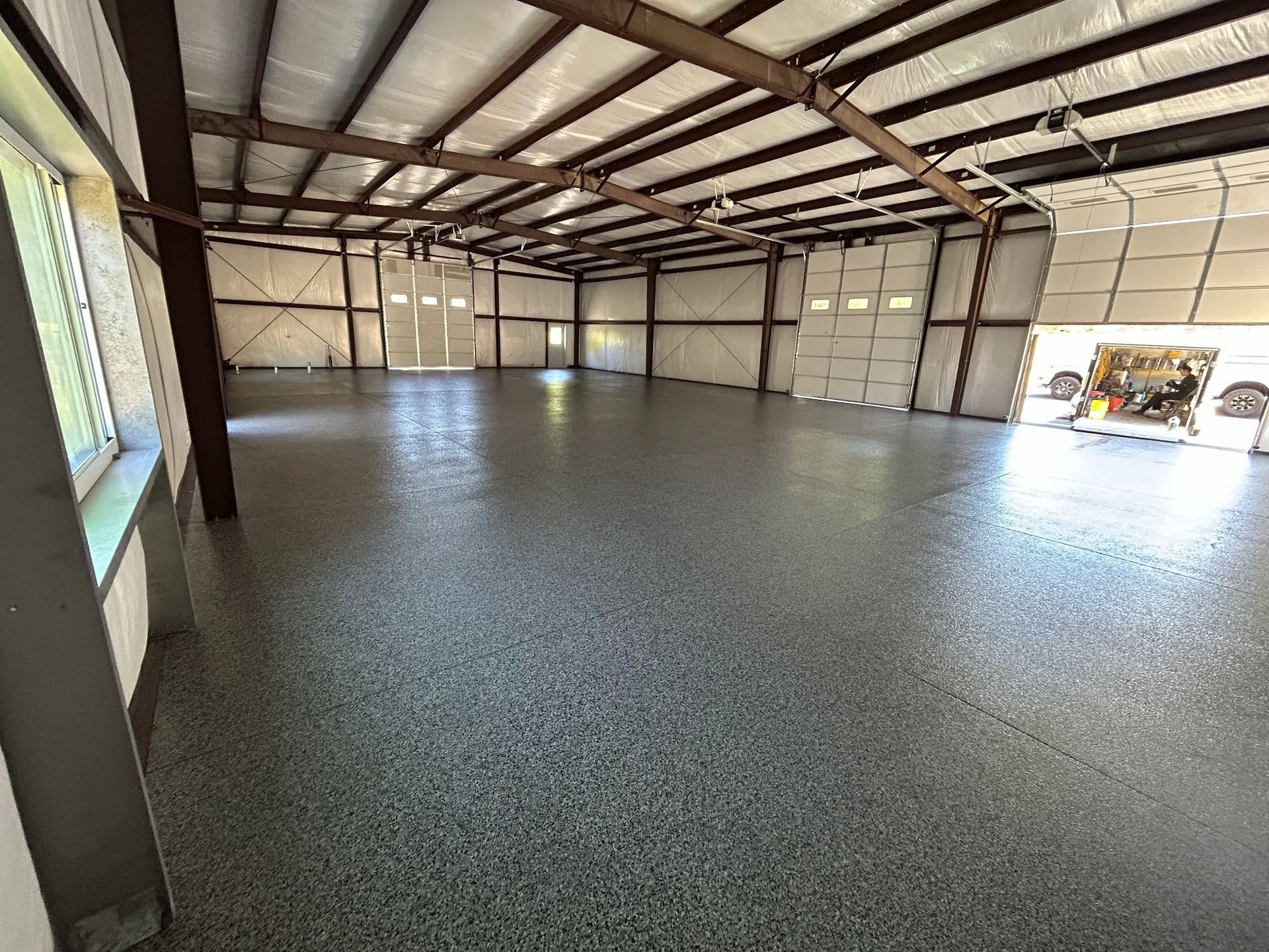 Interior of a large metal building with a polished speckled gray floor, exposed beams, and a window and open garage door.