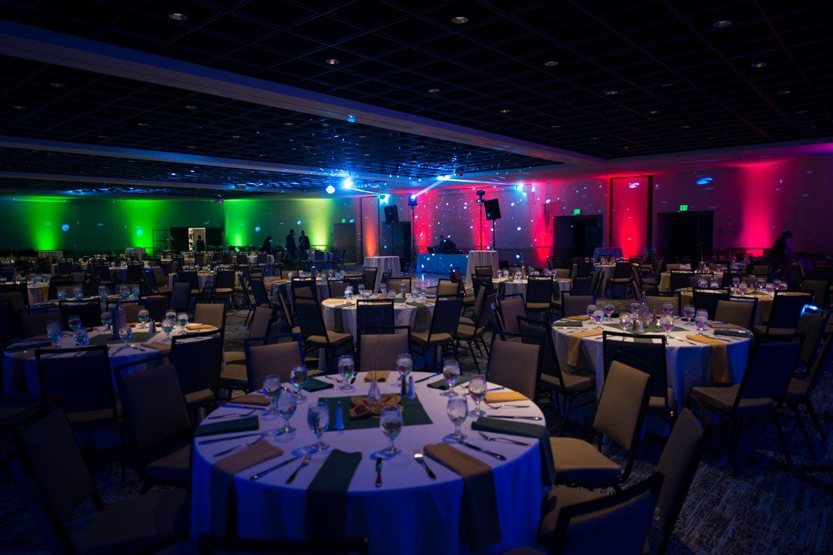 A large banquet hall with round tables set for a formal dinner, lit with green, blue, and red stage lights.