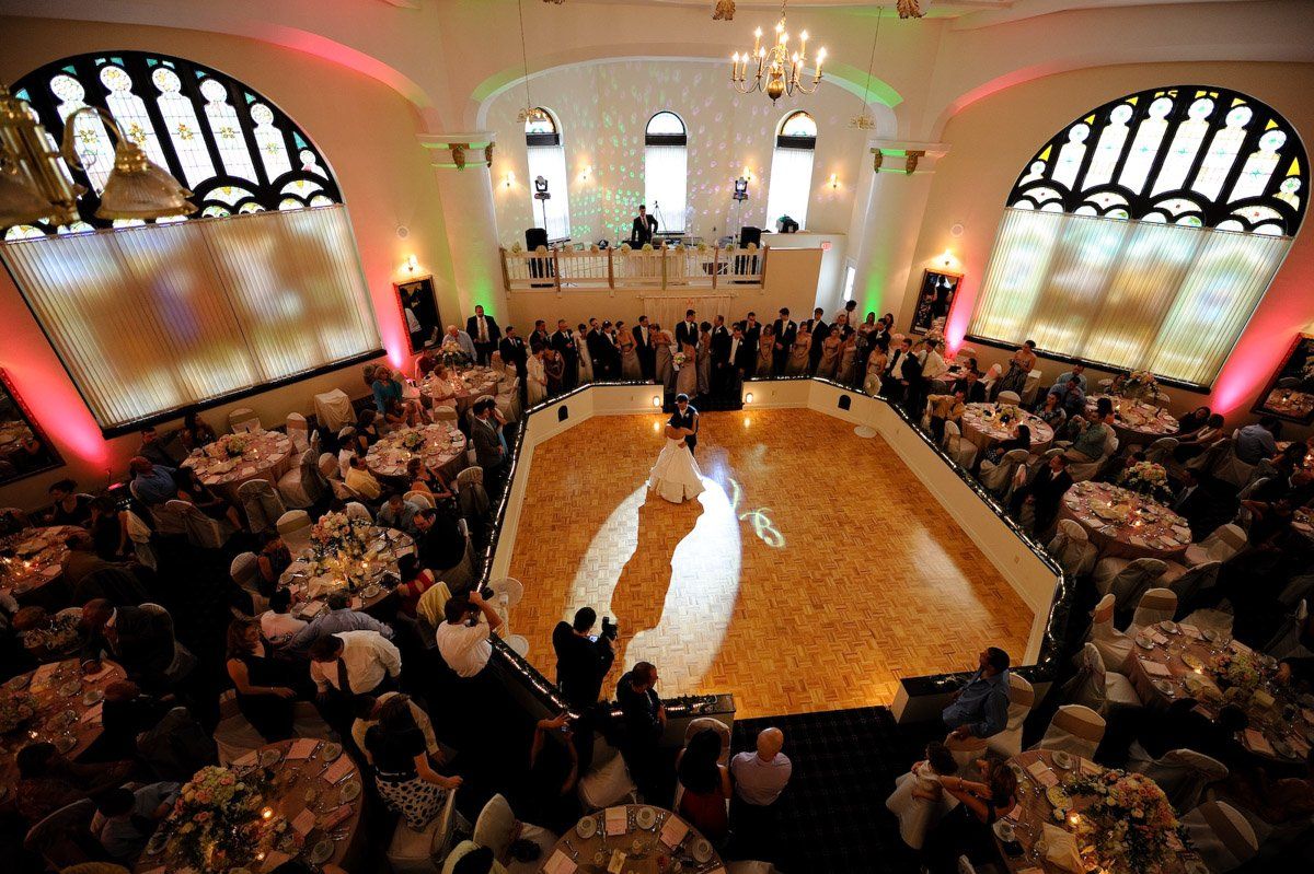 Wedding reception: Bride and groom on dance floor, guests at tables, ornate building interior, ambient lighting.