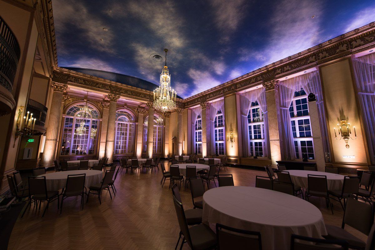 Elegant ballroom with round tables, tall windows, and a cloudy ceiling.
