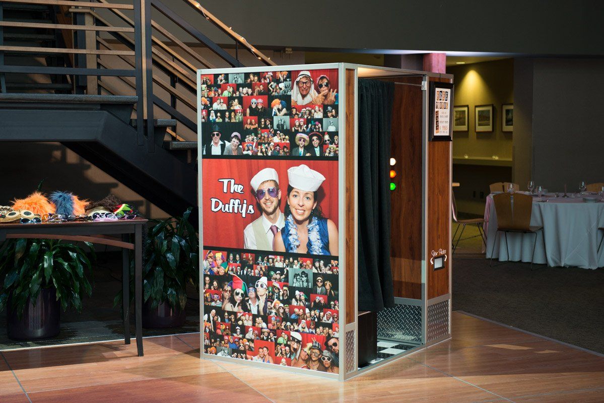 Photo booth with couple collage on the front. Setting is an event space, with a table, stairs, and round tables.