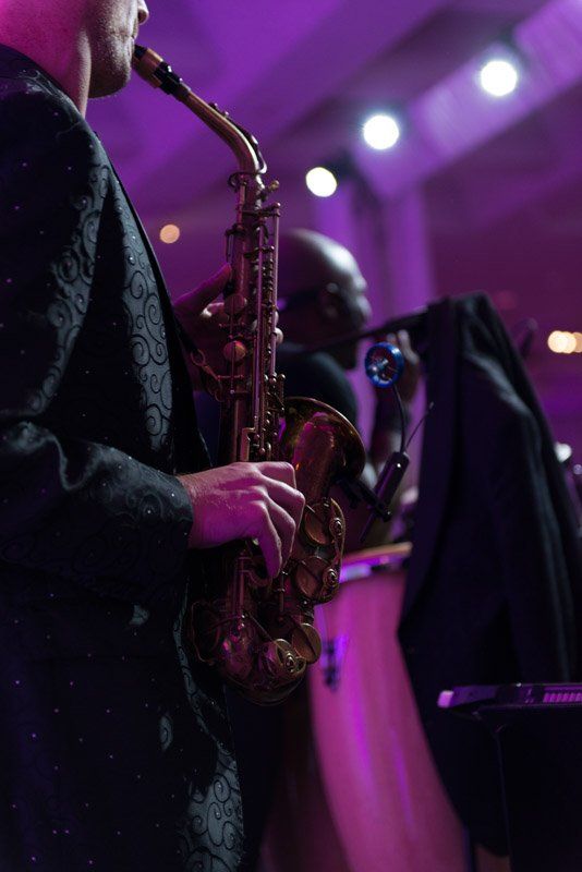 Saxophonist in black suit playing, illuminated by purple stage lights.