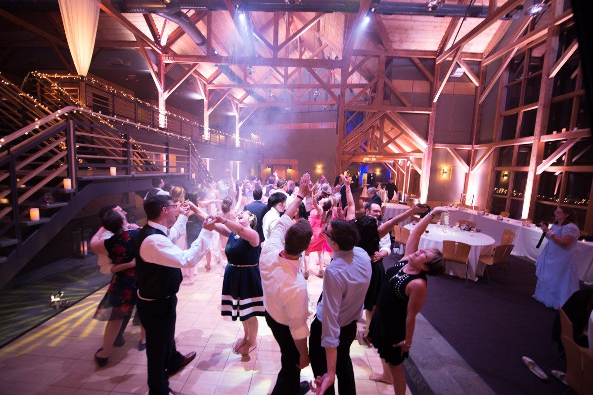 People dancing at a brightly lit wedding reception in a rustic barn setting.