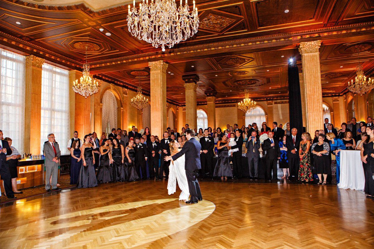 Wedding reception: Bride and groom dance in ballroom, surrounded by guests. Ornate gold decor.