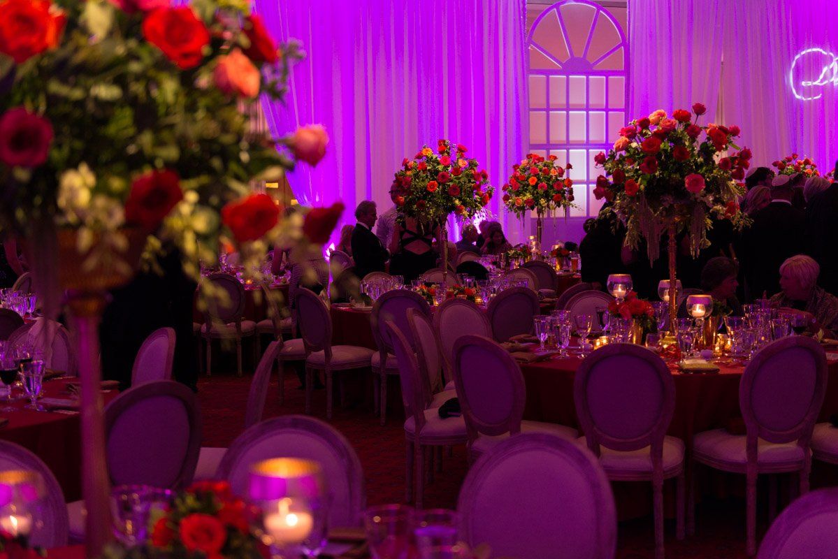 Formal dining room, tables set with red linens and floral centerpieces, bathed in purple light.