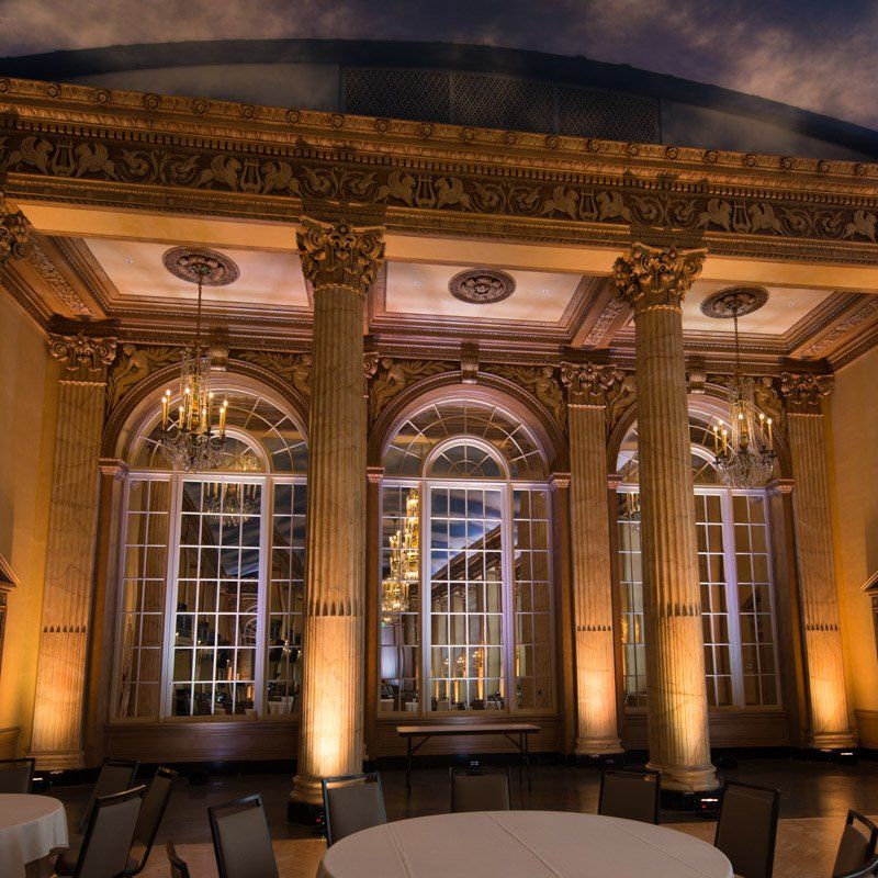 A grand ballroom with ornate gold columns, large windows, chandeliers, and round tables set for a banquet.