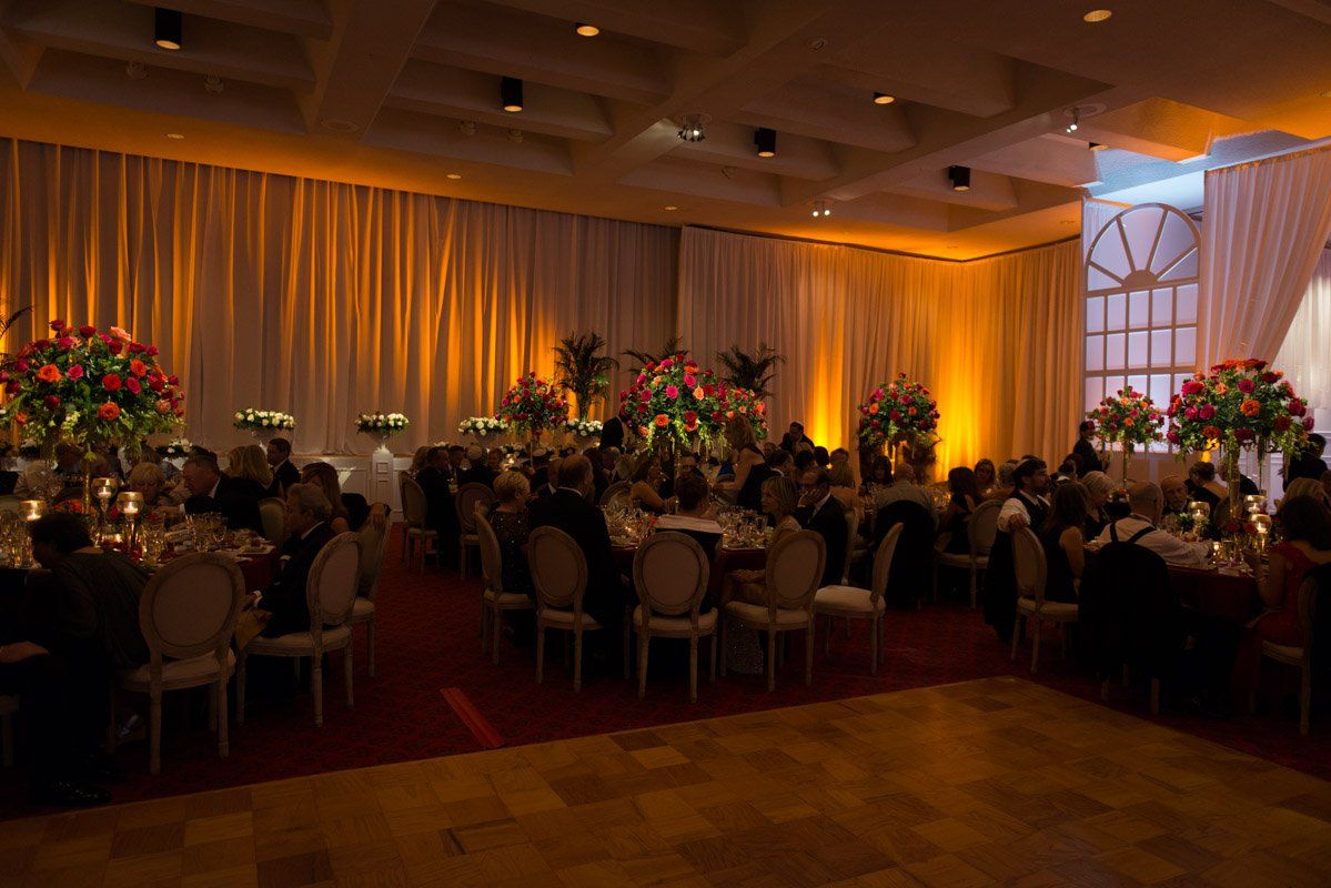 Formal dinner party in a ballroom, warm lighting, guests seated at tables, flowers, and white curtains.