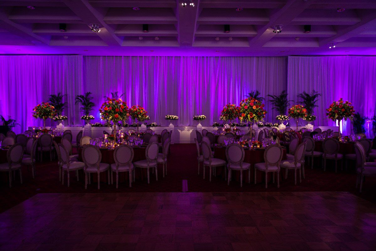 Elegant ballroom set for event, bathed in purple lighting. Tables with floral centerpieces are arranged for dining.