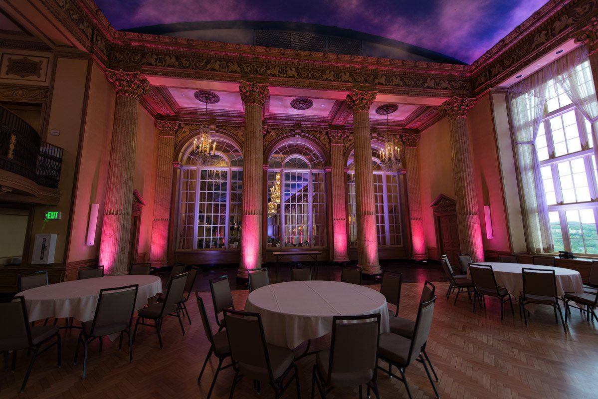 A ballroom with round tables and chairs, accented by pink uplighting and ornate columns, near large windows.