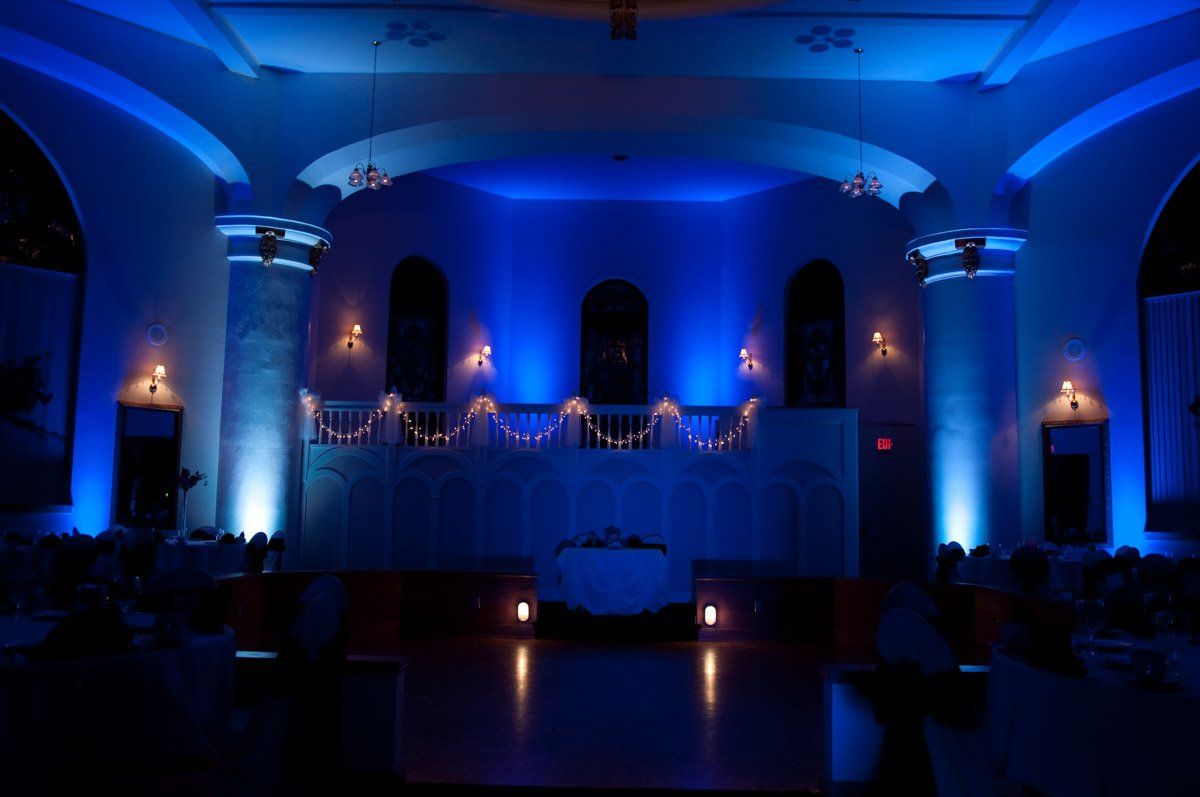 Blue-lit ballroom with ornate architecture. Empty stage, tables set up in audience, creating a sophisticated ambiance.