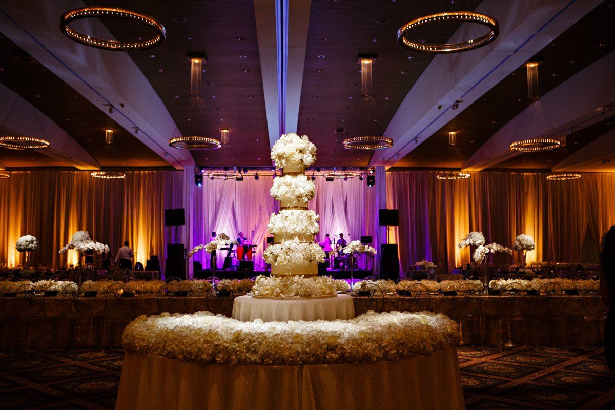 Elegant wedding reception with a tiered cake in a ballroom, bathed in warm and purple lighting.