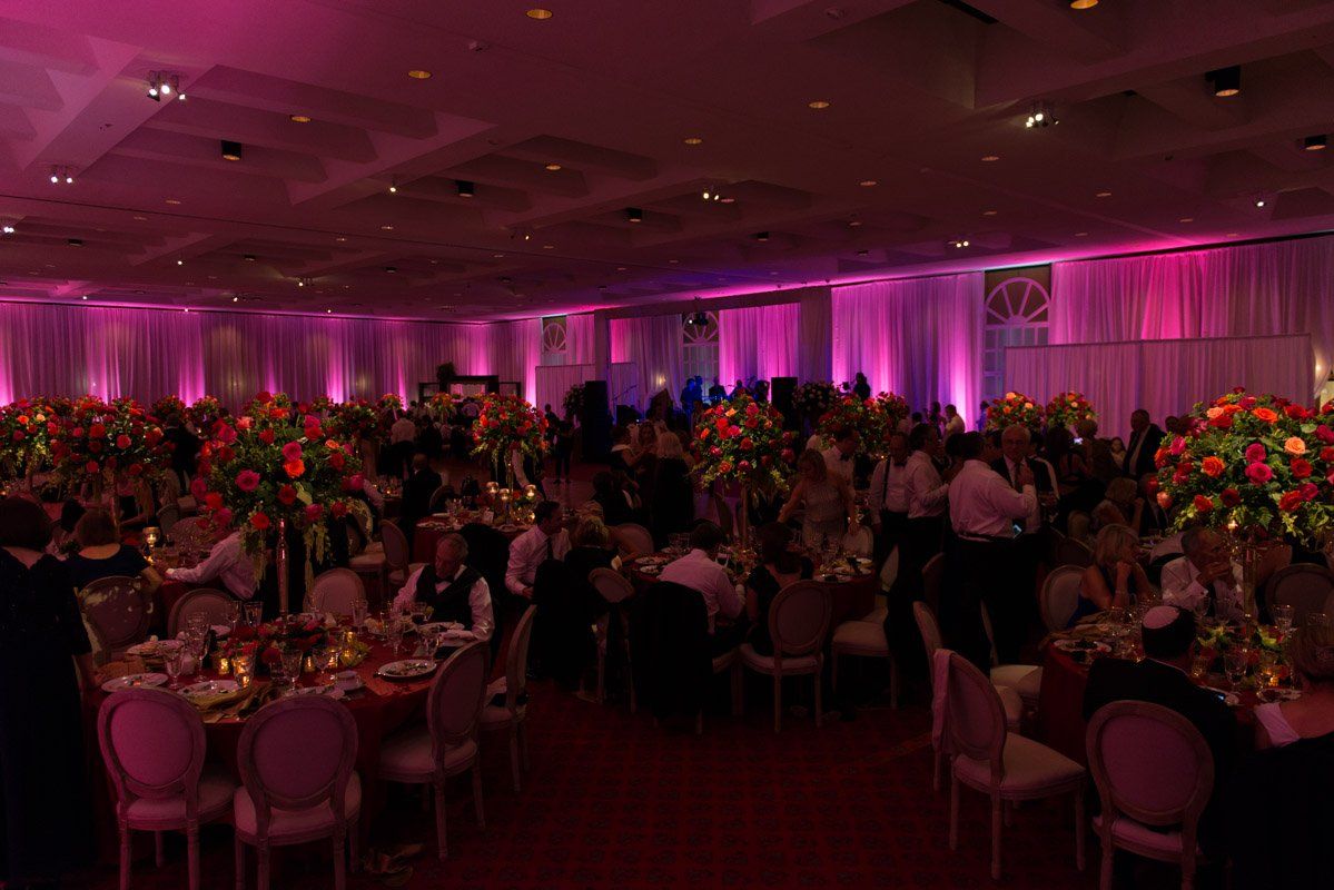 A ballroom set for a gala, featuring people at tables with tall floral centerpieces, illuminated with pink lighting.
