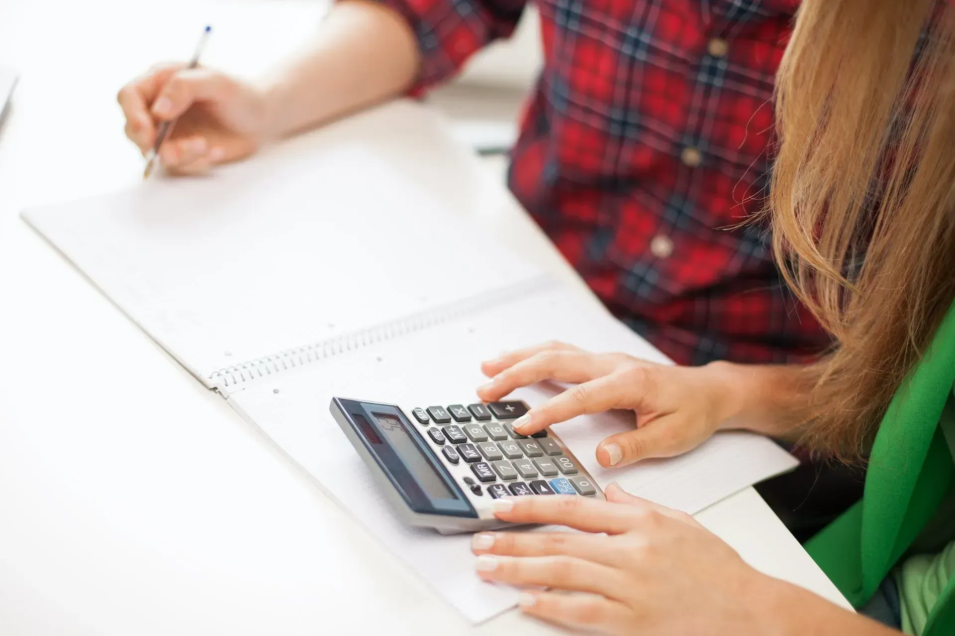 A woman is using a calculator while a man is writing in a notebook.