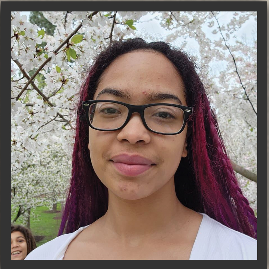 A woman with purple hair and glasses is standing in front of a cherry blossom tree.