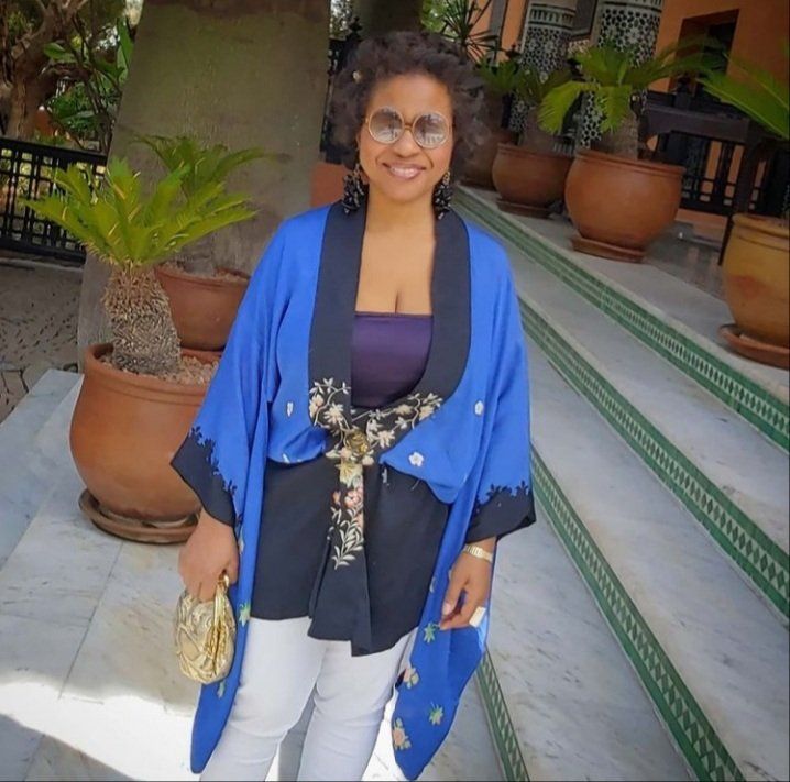 A woman wearing a blue and black kimono stands in front of potted plants
