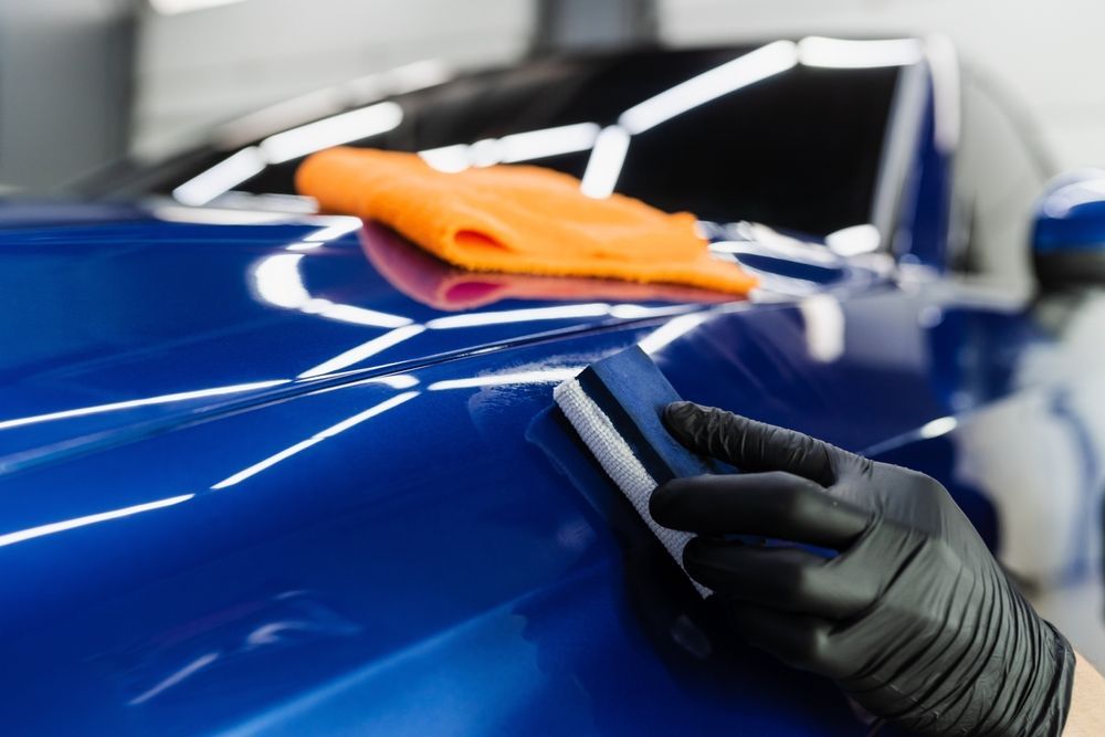 Gloved hand wiping a blue car with an orange microfiber cloth in a garage