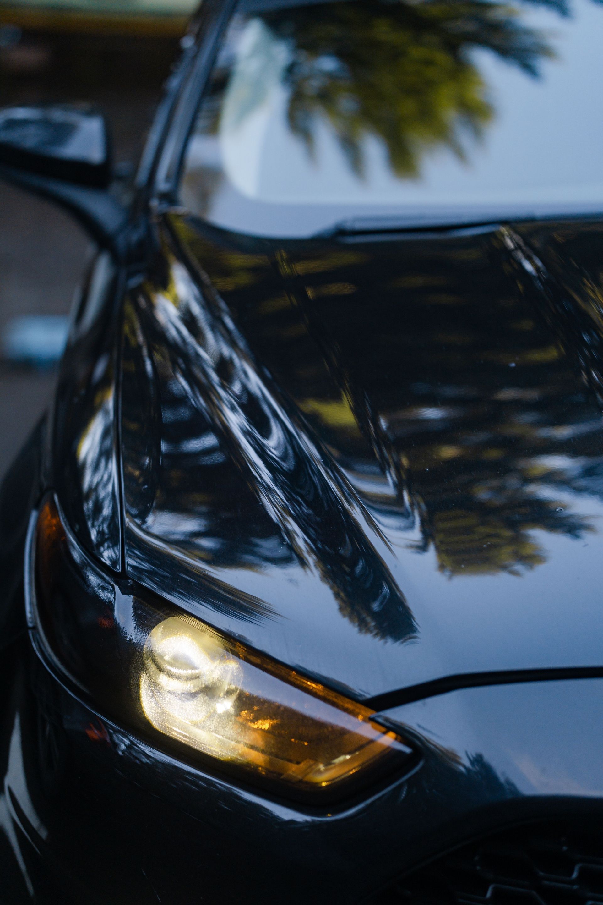 Close-up of a black car’s front corner with a lit headlight and reflections of trees on the hood