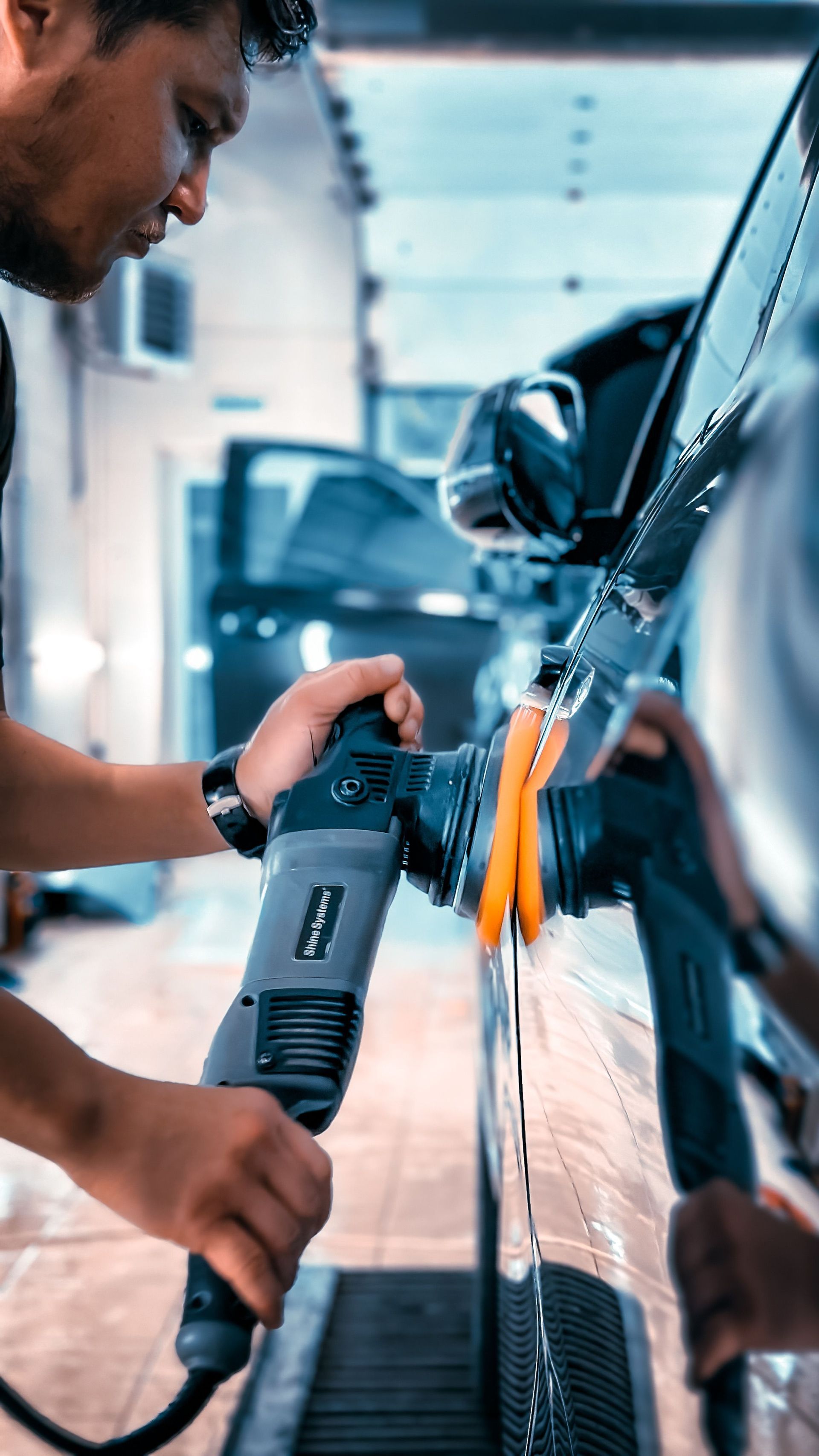 Man polishing a black car door with an electric buffer in a garage