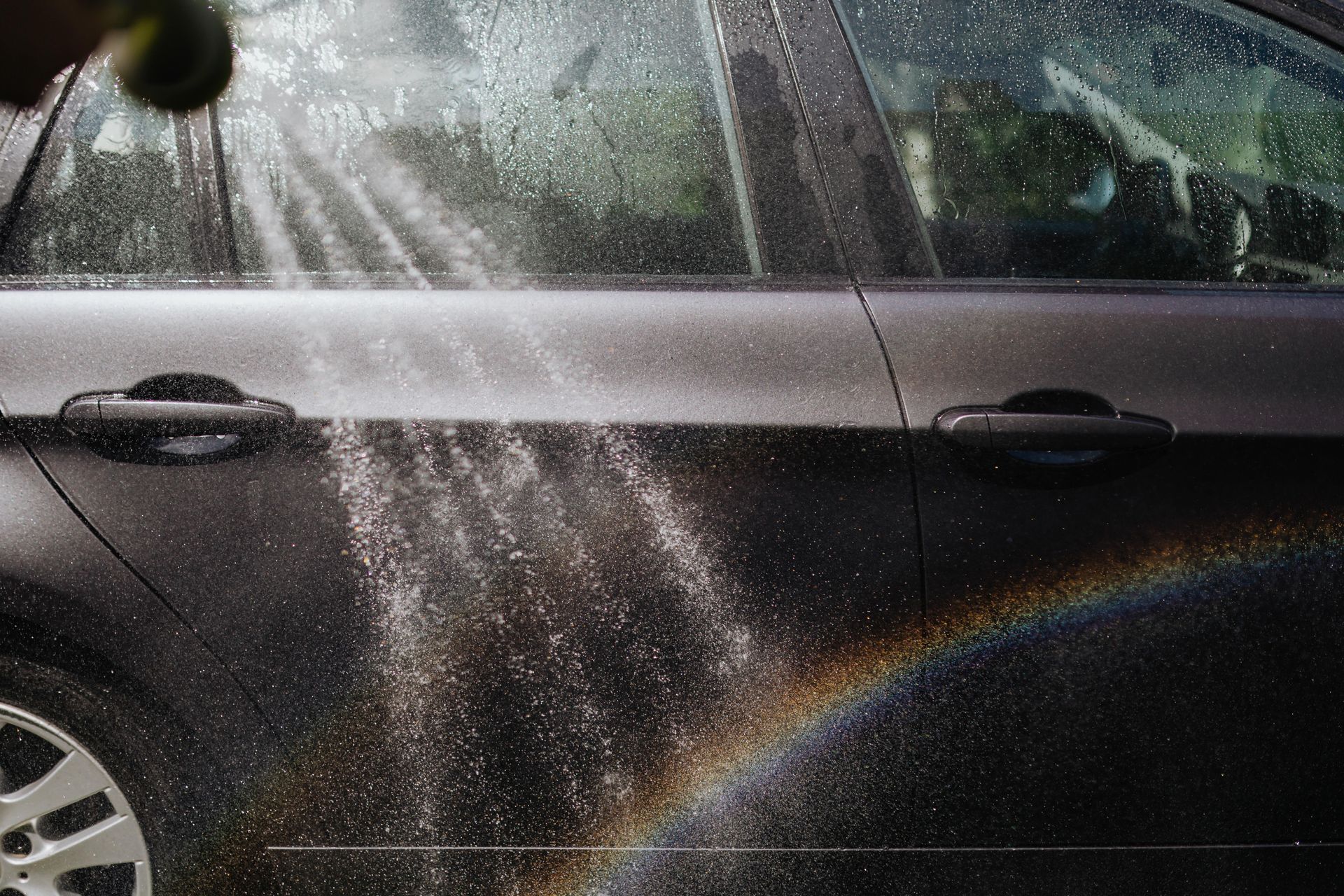 Black car door and window being sprayed with water, with a rainbow arc across the lower half.
