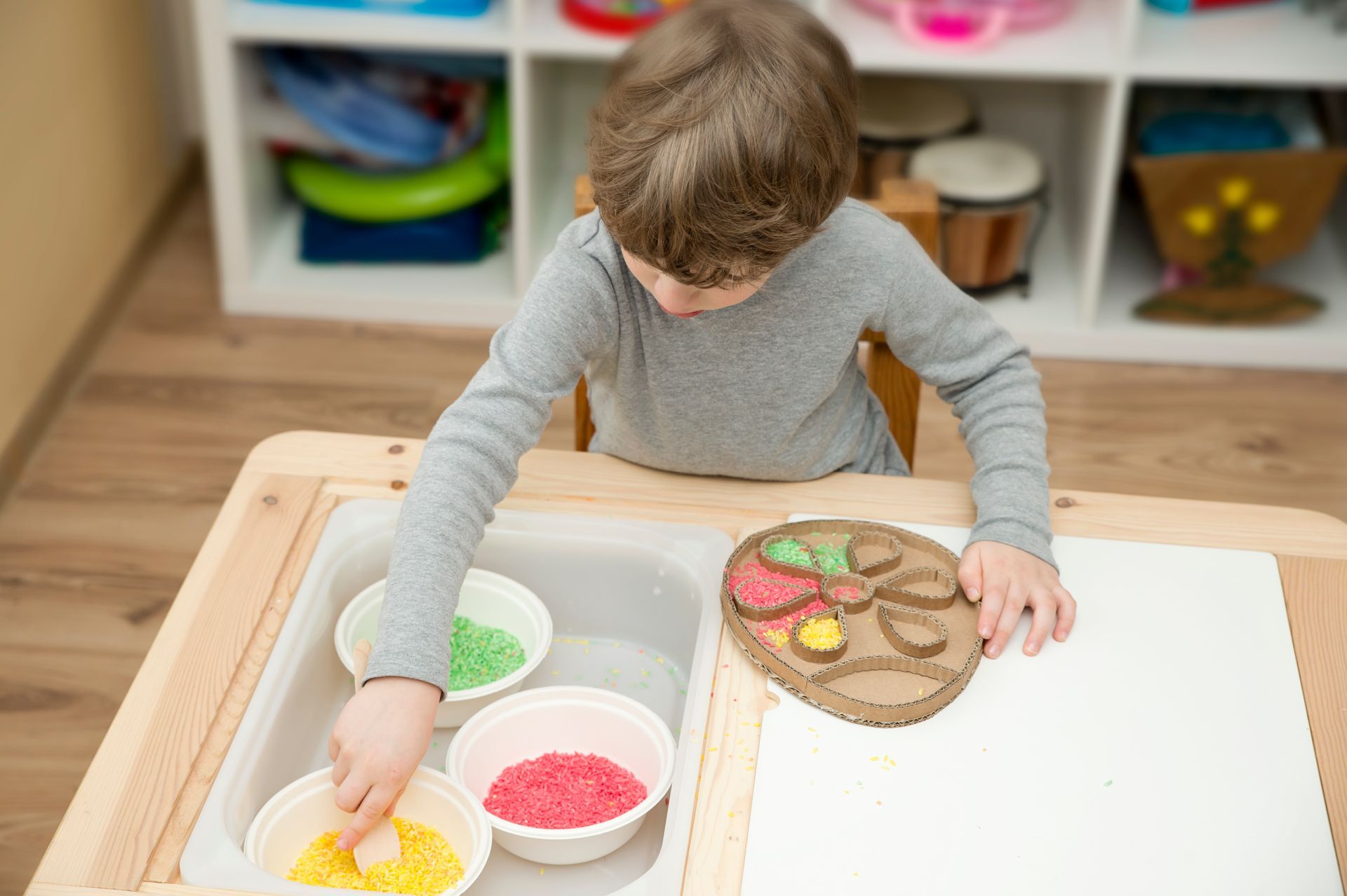 Boy at a table with colored sand, placing it on a cardboard shape. Boy at a table with colored sand, placing it on a cardboard shape.