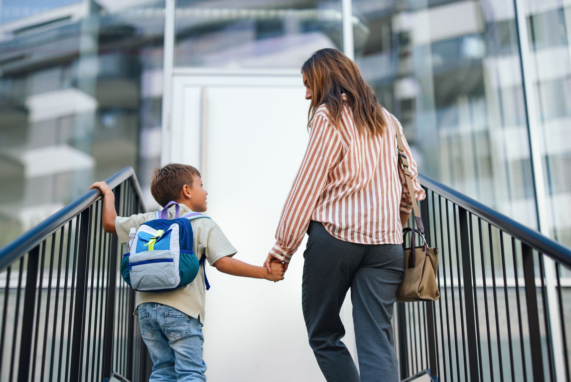Child with backpack holding adult's hand walking up stairs to building.