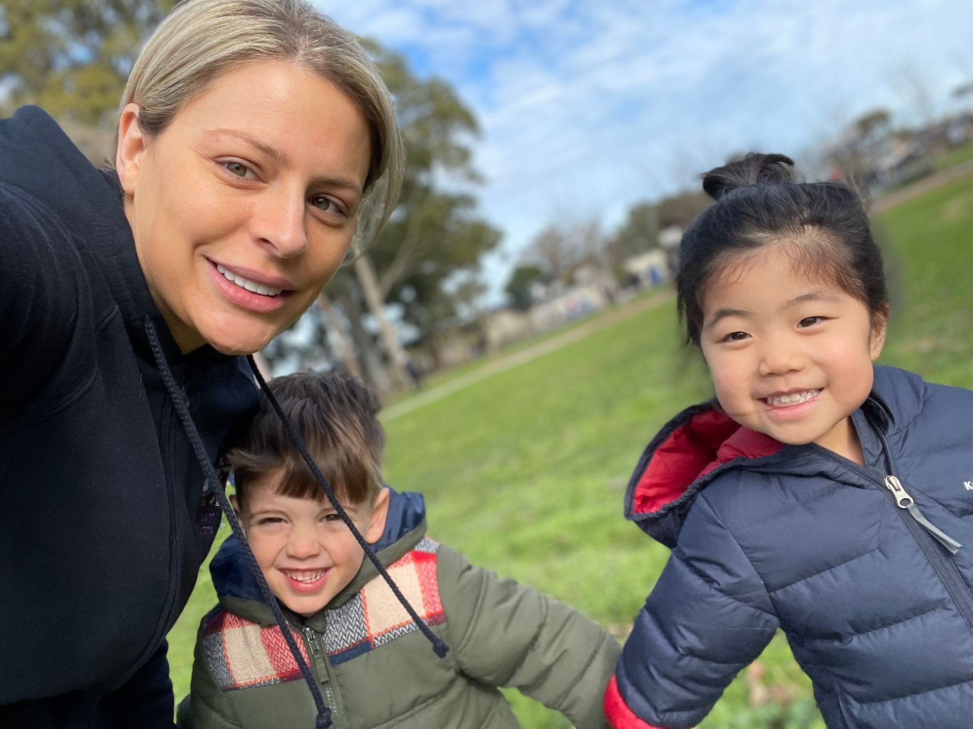 Woman and two children smiling outdoors on a sunny day. Green grass, trees, blue jackets.